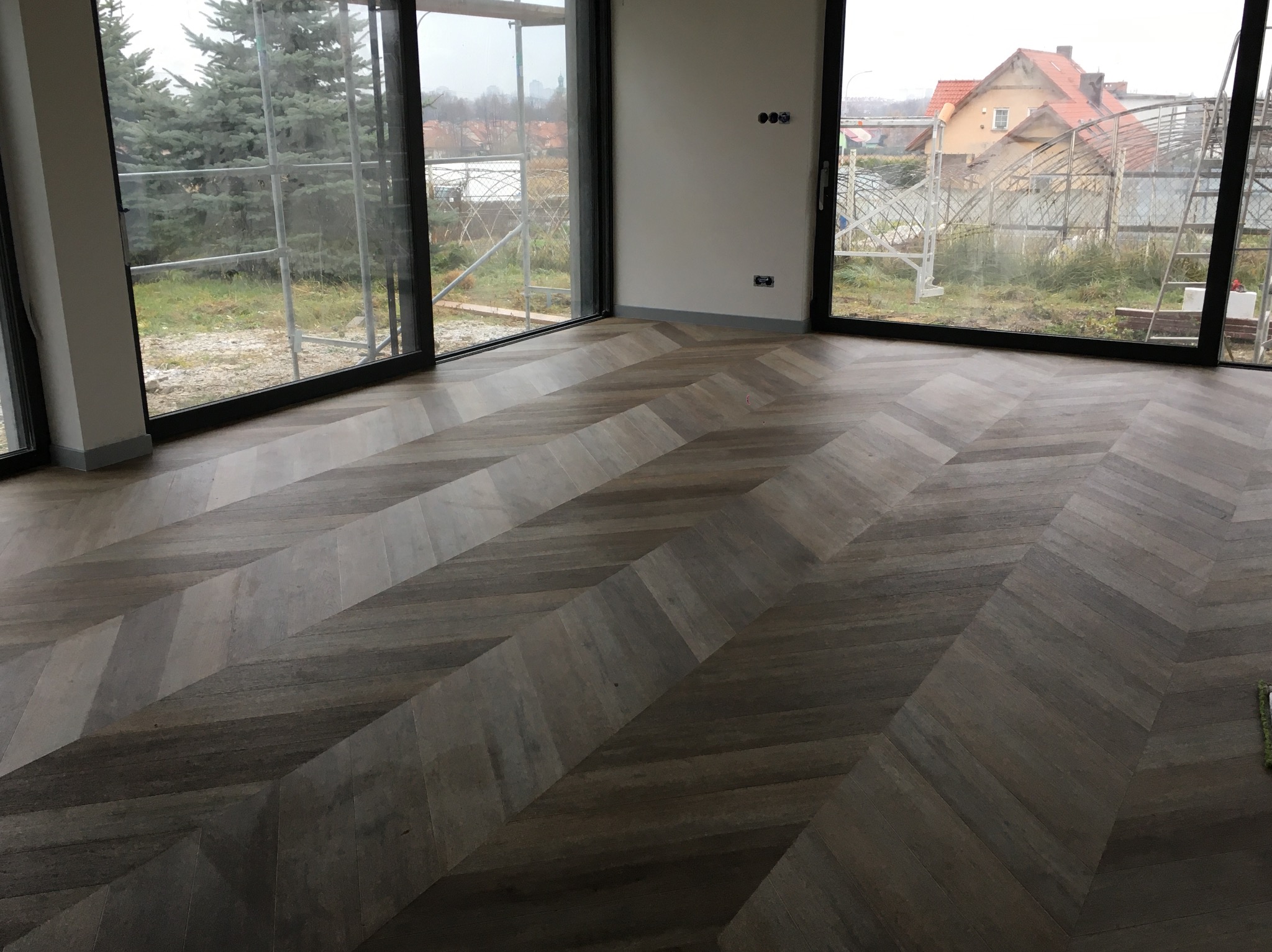 Ułożony parkiet w jodełkę w nowym domu z dużymi oknami, widok na ogród i budynek w tle.