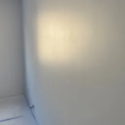 Świeżo pomalowane wnętrze pokoju w odcieniach szarości, widoczne delikatne refleksy światła na gładkiej ścianie i fragment zabezpieczonej taśmą malarską podłogi.