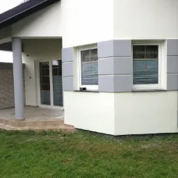 Wykończona elewacja domu jednorodzinnego w jasnych kolorach z szarymi detalami wokół okien i kolumną podtrzymującą zadaszenie wejścia, widok z poziomu trawnika.