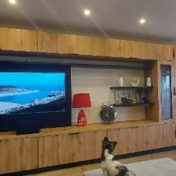 Zabudowa meblowa w salonie z telewizorem, półkami i przeszkloną witryną, wykonana z drewna w ciepłym odcieniu, z widoczną lampą z czerwonym abażurem i zegarem oraz psem rasy yorkshire terrier...