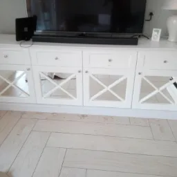 Biała szafka RTV z frezowanymi drzwiczkami z lustrami, ustawiona na podłodze z jasnych paneli ułożonych w jodełkę, z telewizorem i lampą na blacie.