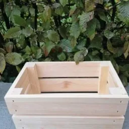 Surowa, jasna, drewniana skrzynka z desek na tle zielonych liści, ustawiona na szarej, betonowej powierzchni.