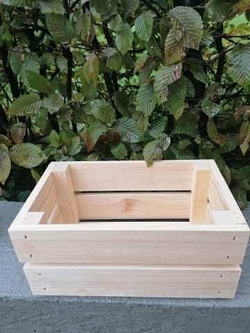 Surowa, jasna, drewniana skrzynka z desek na tle zielonych liści, ustawiona na szarej, betonowej powierzchni.