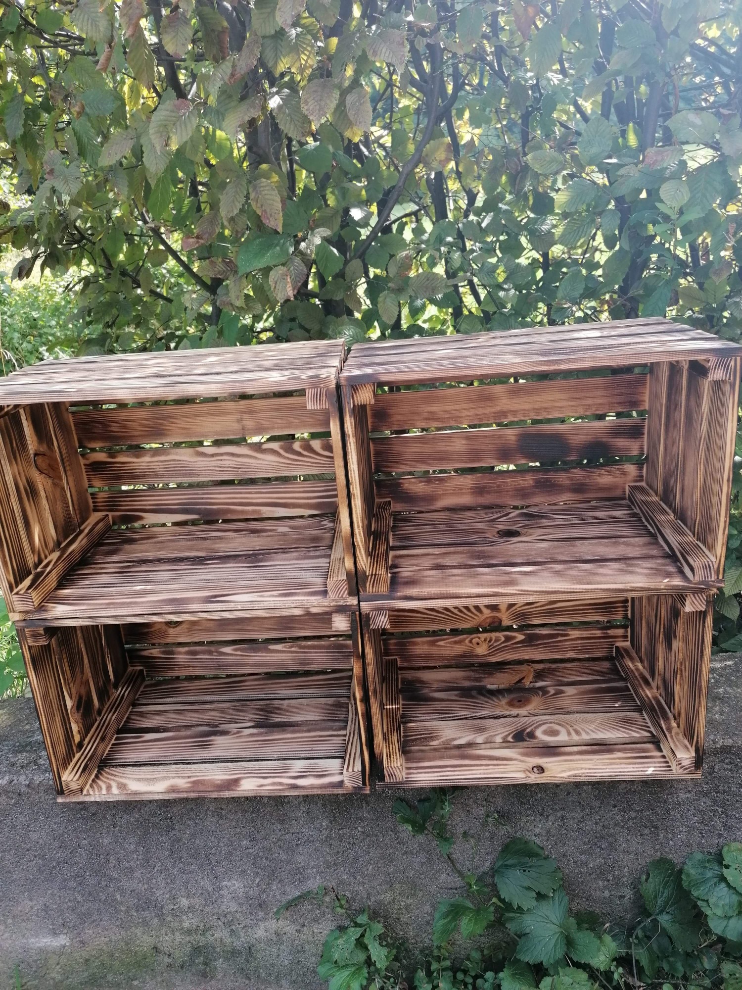 Dwa rzędy wypalanych, drewnianych skrzynek ustawionych na betonowej powierzchni, na tle zieleni.