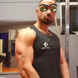 Mężczyzna w czarnej koszulce CONVERSE prezentuje swoją muskulaturę w siłowni, z naklejkami na twarzy.