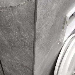 Szare płytki imitujące kamień na ścianie w łazience, widoczny fragment toalety, fuga w kolorze płytek.