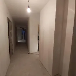 Korytarz w trakcie prac wykończeniowych; ściany pokryte gładzią, surowa żarówka zwisa z sufitu, widoczne ślady prac na podłodze.