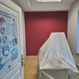 Pokój biurowy z pomalowaną na bordowo ścianą, zabezpieczonym meblem folią malarską i plakatem na drzwiach.