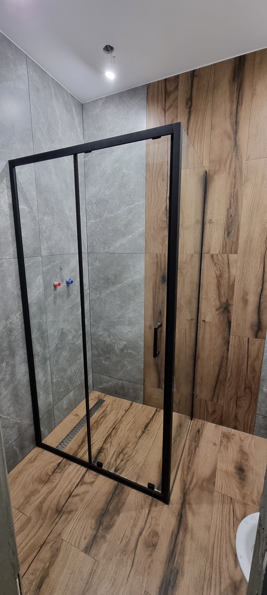 Nowoczesna kabina prysznicowa z czarnymi profilami, szklanymi drzwiami przesuwnymi, zamontowana w łazience wykończonej płytkami imitującymi drewno i szary marmur, z widocznymi elementami instalacji...