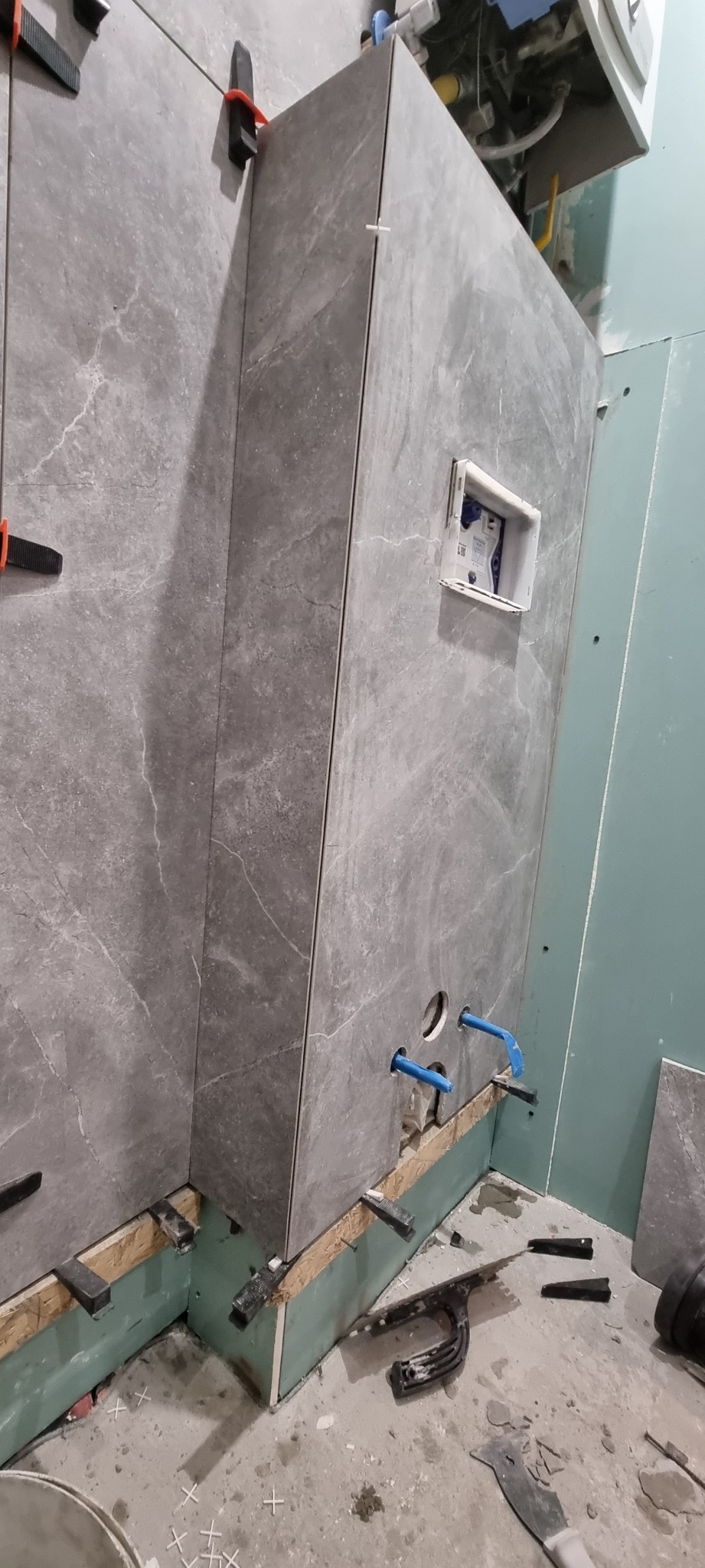 Montaż płytek imitujących marmur w łazience, widoczne poziomice, krzyżyki dystansowe i otwory na instalacje sanitarne, ściana z płyty gipsowo-kartonowej w tle.