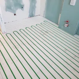 Zielone rurki ogrzewania podłogowego ułożone na białej izolacji w surowym wnętrzu z płytami gipsowo-kartonowymi na ścianach, widoczne przyłącza instalacji.