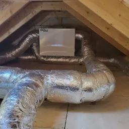 Rekuperator marki WANAS zamontowany na poddaszu, widoczne izolowane przewody wentylacyjne w srebrnej folii aluminiowej oraz drewniana konstrukcja dachu.
