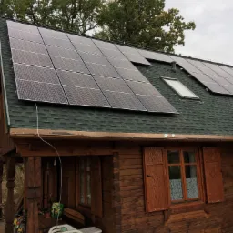 Drewniany dom z zielonym dachem krytym gontem, na którym zamontowano panele fotowoltaiczne i okna dachowe.