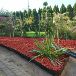 Rabata z czerwoną korą, obrzeżem z metalu i młodymi drzewkami, widok z boku, trawnik w tle.