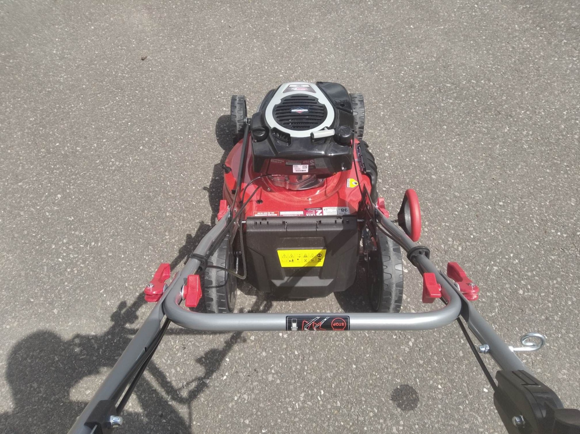 Czerwona kosiarka spalinowa marki Briggs & Stratton, widok z góry na asfaltowym podłożu, z widocznym cieniem osoby obsługującej.