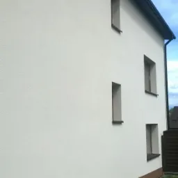 Elewacja budynku po tynkowaniu, z widocznymi oknami i brązowym cokołem. Tekstura tynku baranek w jasnym odcieniu, czysta i schludna.
