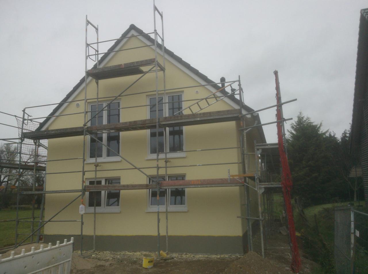 Żółty dom jednorodzinny w trakcie tynkowania elewacji, widoczne rusztowania i niewielki fragment sąsiedniego budynku po prawej stronie.