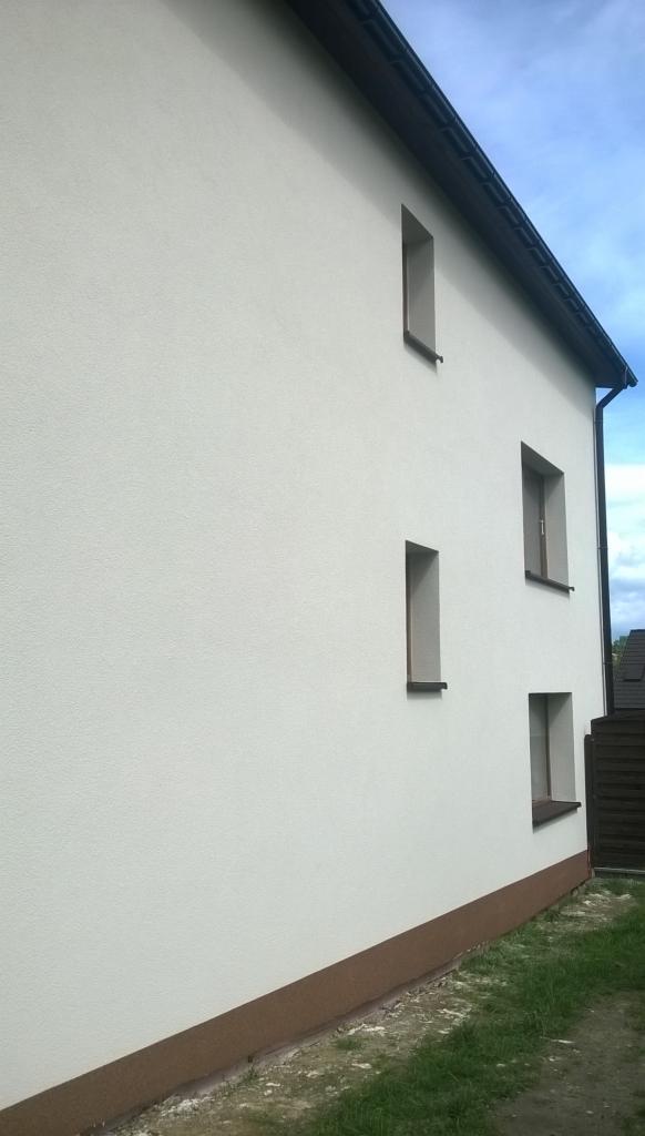 Elewacja budynku po tynkowaniu, z widocznymi oknami i brązowym cokołem. Tekstura tynku baranek w jasnym odcieniu, czysta i schludna.