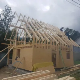 Wznoszenie konstrukcji domu z drewnianych belek i płyt OSB, widoczni pracownicy montujący więźbę dachową na tle zalesionych wzgórz i pochmurnego nieba.