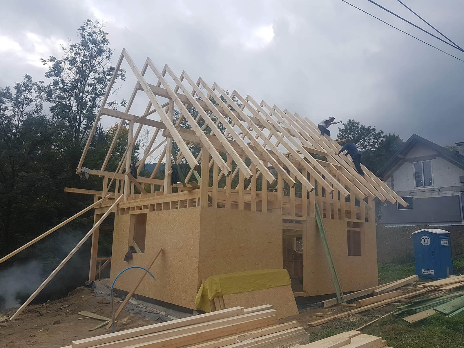 Wznoszenie konstrukcji domu z drewnianych belek i płyt OSB, widoczni pracownicy montujący więźbę dachową na tle zalesionych wzgórz i pochmurnego nieba.