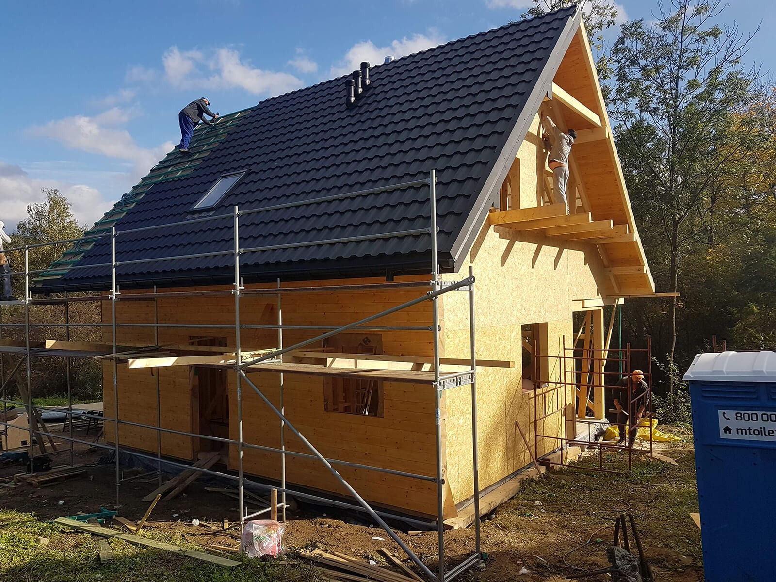 Wznoszenie domu z drewnianą konstrukcją szkieletową: robotnicy montują czarną blachodachówkę na dachu, widoczne rusztowania i częściowo obłożone ściany z płyt OSB, przenośna toaleta na pierwszym...