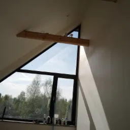 Ściana pod skosem z oknem trójkątnym, widoczne ślady po szpachlowaniu i drewniana belka pod sufitem.