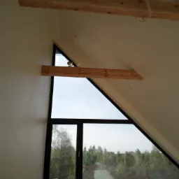 Poddasze z oknem o nietypowym trójkątnym kształcie, świeżo pomalowane ściany w odcieniach bieli, drewniane belki stropowe