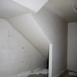Surowe wnętrze poddasza w trakcie wykańczania: ściany i skośny sufit po wstępnym przygotowaniu, widoczne kable elektryczne i niedokładności na podłodze.