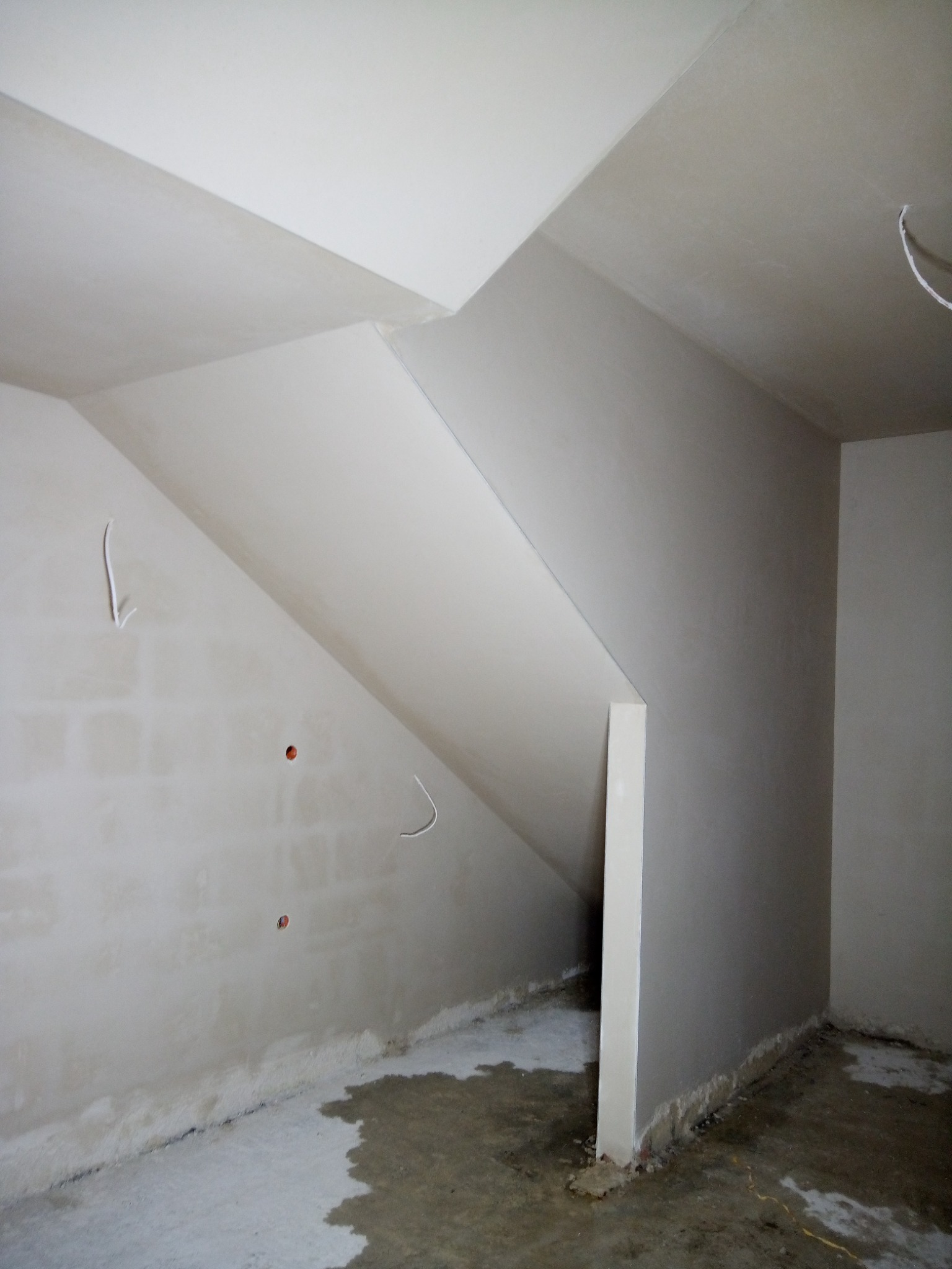 Surowe wnętrze poddasza w trakcie wykańczania: ściany i skośny sufit po wstępnym przygotowaniu, widoczne kable elektryczne i niedokładności na podłodze.