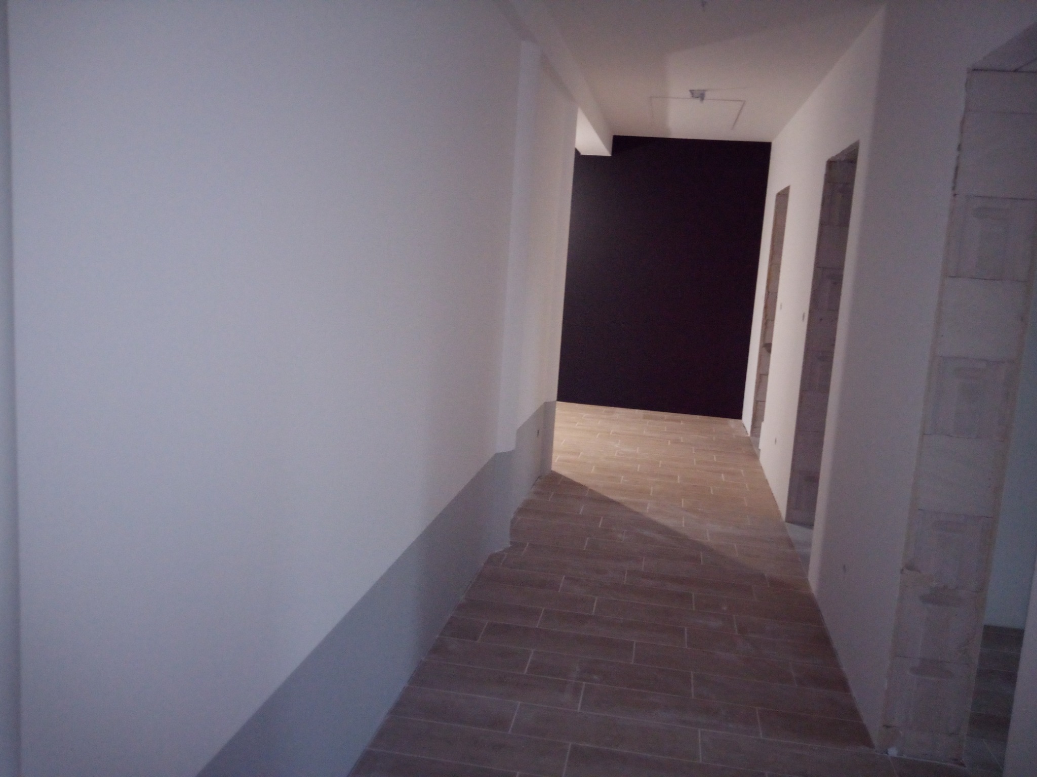 Korytarz w trakcie prac wykończeniowych: ściany częściowo pomalowane na biało, z szarym pasem przy podłodze; podłoga wyłożona brązowymi płytkami imitującymi drewno; widoczne surowe wnęki drzwiowe...