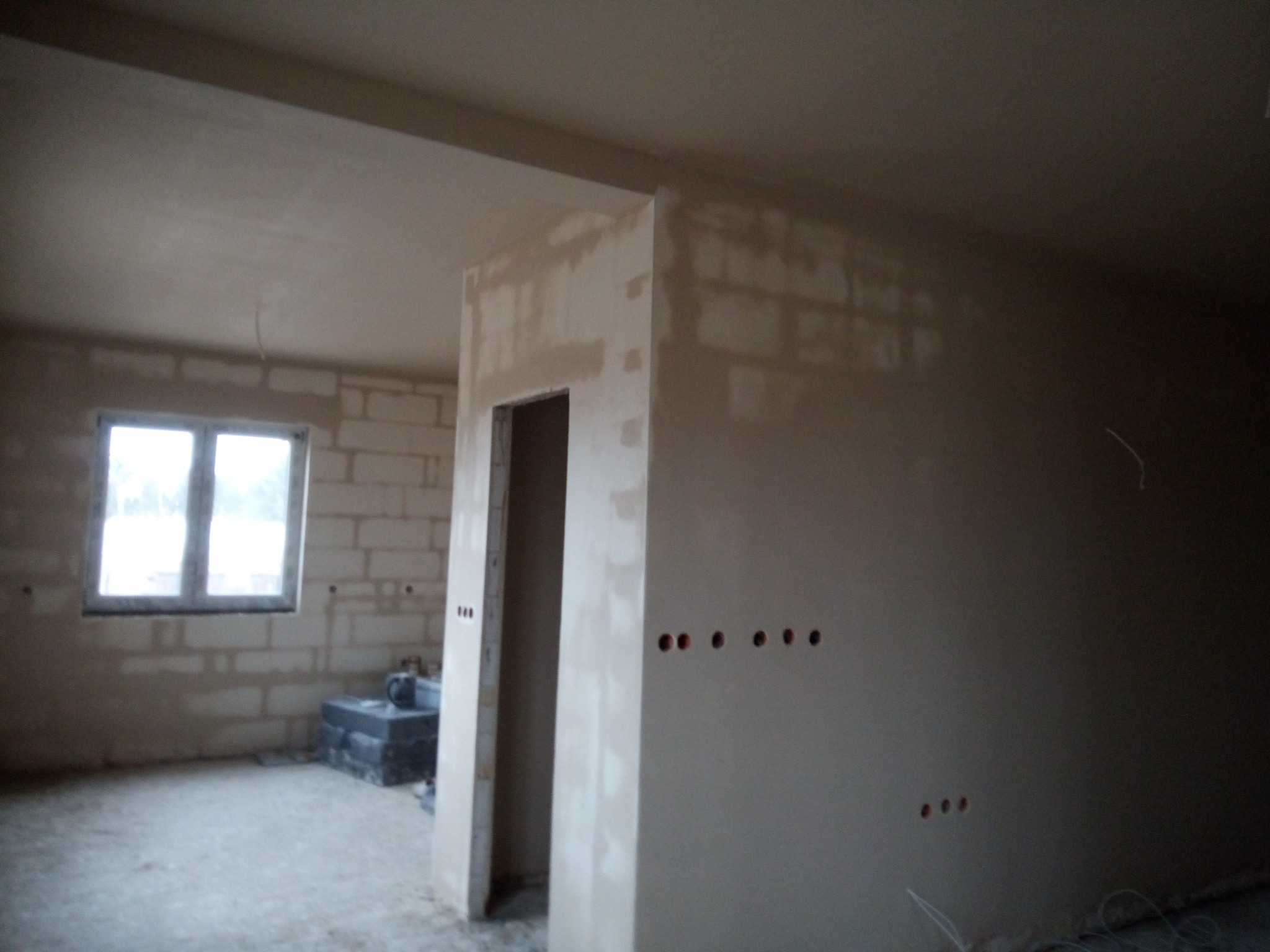 Wnętrze w trakcie prac wykończeniowych: ściany i sufit częściowo pokryte zaprawą, widoczne otwory na gniazdka elektryczne, bloczki betonowe i okno w tle.