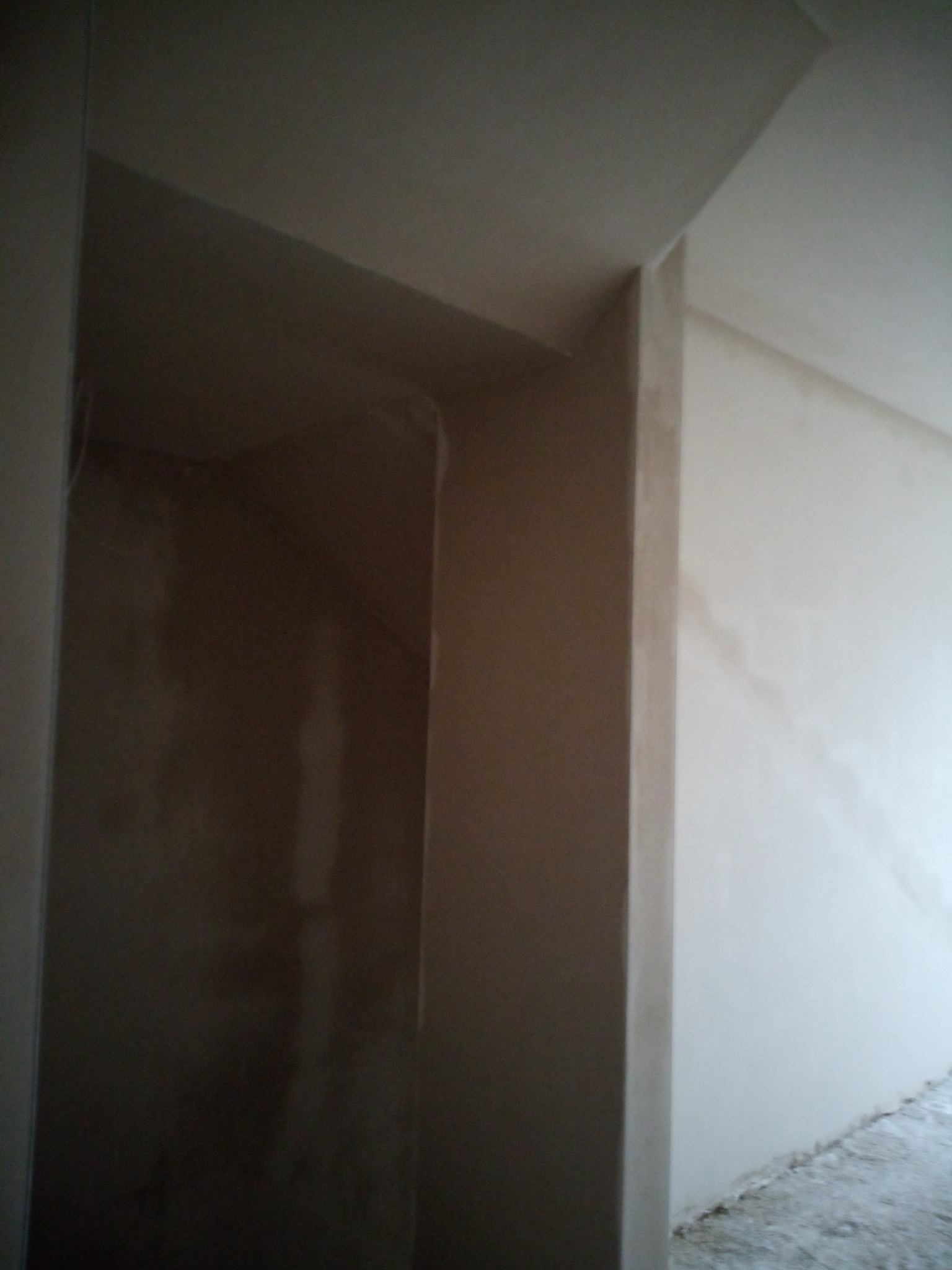 Wnętrze w trakcie prac wykończeniowych, widoczna surowa ściana z nałożoną warstwą szpachli, częściowo oświetlona, z widocznymi niedoskonałościami powierzchni.