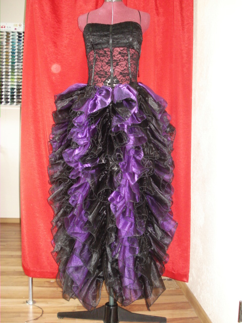 Czarno-fioletowa suknia balowa z falbanami na manekinie krawieckim przed czerwoną zasłoną, widoczna szpula nici w tle.