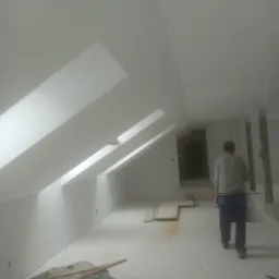 Poddasze po pracach wykończeniowych, widoczne skosy z oknami dachowymi, pomalowane na biało, na podłodze leżą narzędzia i płyty gipsowe, w tle sylwetka mężczyzny.