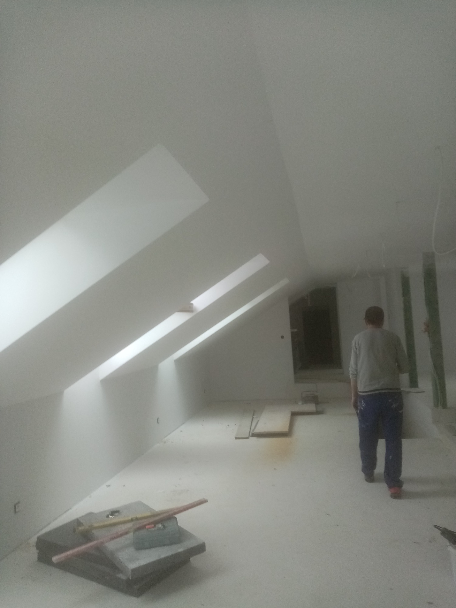 Poddasze po pracach wykończeniowych, widoczne skosy z oknami dachowymi, pomalowane na biało, na podłodze leżą narzędzia i płyty gipsowe, w tle sylwetka mężczyzny.