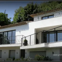 Elegancki, biały dom z czarnymi, poziomymi balustradami. Ujęcie z dołu, eksponujące balkony i okna. Tło stanowią drzewa i krzewy. Widoczny adres strony internetowej.