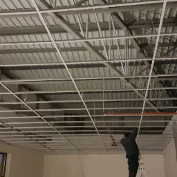 Montaż metalowej konstrukcji sufitu podwieszanego w pomieszczeniu biurowym, widoczny pracownik na drabinie i laserowy niwelator.