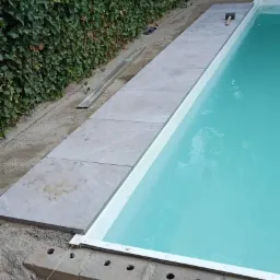 Etap budowy basenu: układanie jasnoszarych płyt kamiennych wokół niecki, widoczny mur z pustaków, narzędzia (młotek) i odpływ liniowy, tło stanowi zielony żywopłot.