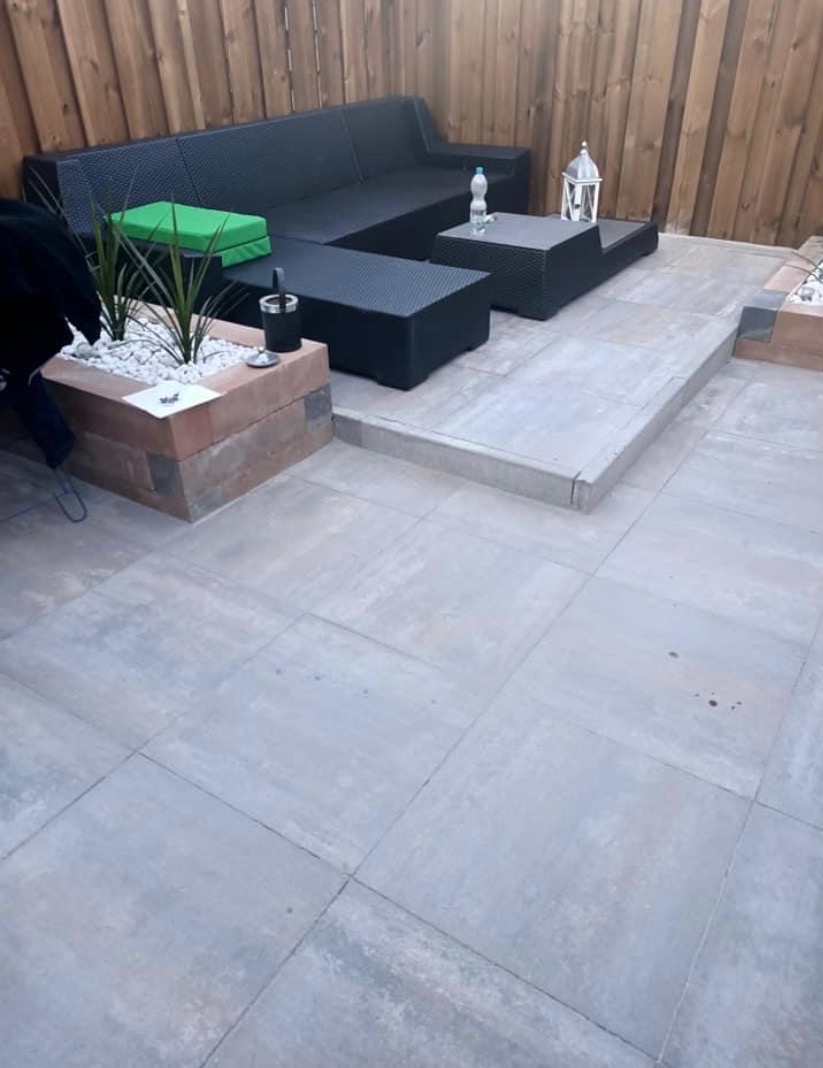 Szare płytki tarasowe tworzące patio z czarnym rattanowym zestawem mebli ogrodowych, podwyższony kwietnik z cegły i drewniane ogrodzenie w tle.