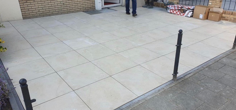 Jasne, kwadratowe płytki tarasowe ułożone na zewnątrz budynku z widocznymi czarnymi słupkami ogrodzeniowymi i fragmentem chodnika z kostki brukowej. W tle widoczne materiały budowlane i fragment...