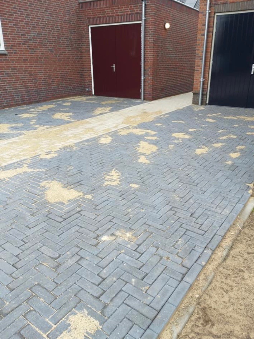 Ułożona szara kostka brukowa w jodełkę na podjeździe, z piaskiem w spoinach, widoczne dwie bramy garażowe w tle.