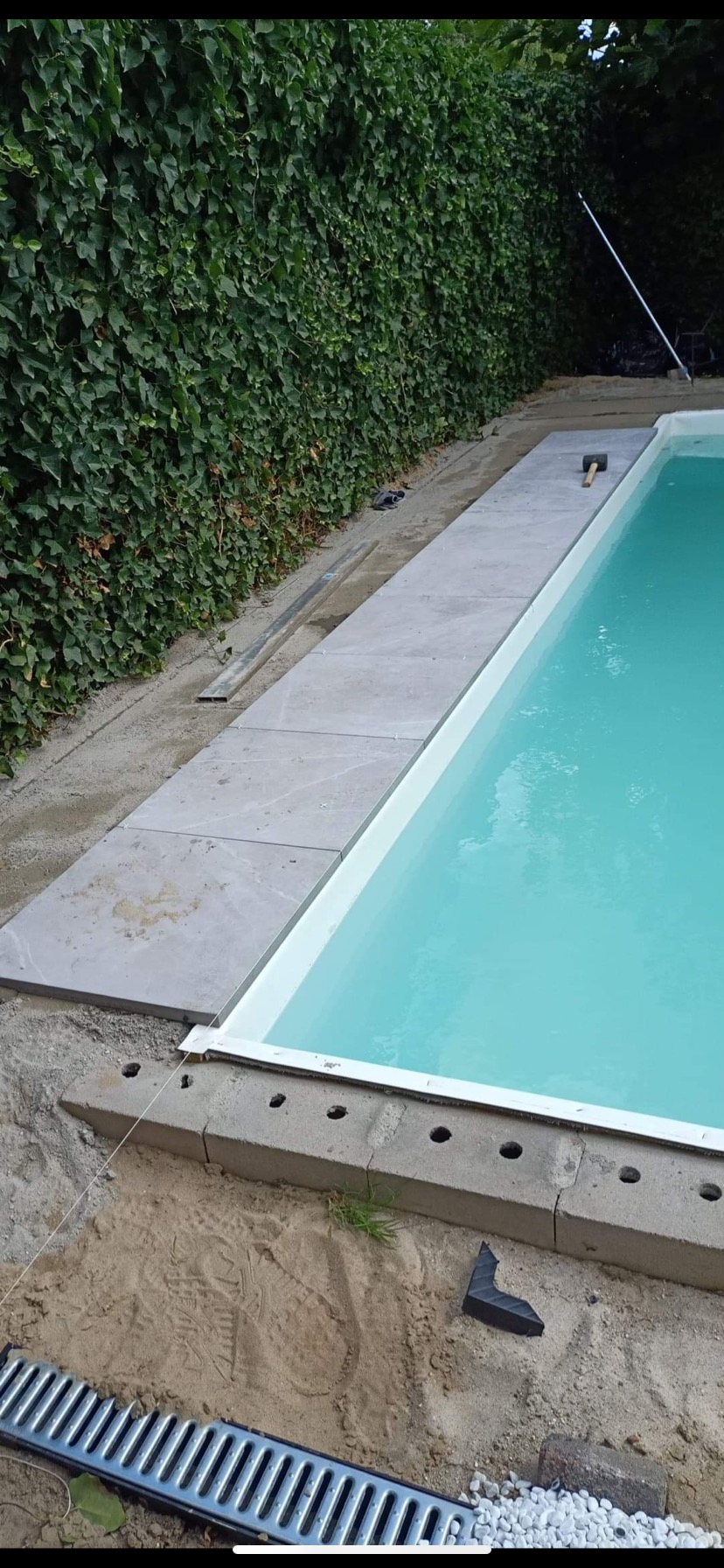 Etap budowy basenu: układanie jasnoszarych płyt kamiennych wokół niecki, widoczny mur z pustaków, narzędzia (młotek) i odpływ liniowy, tło stanowi zielony żywopłot.