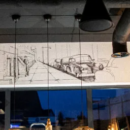 Szkic ołówkiem na ścianie przedstawiający ulicę z zaparkowanymi klasycznymi samochodami i postacią w kapeluszu, widoczny przez okno z odbiciami i elementami wystroju wnętrza, oświetlony wiszącymi...