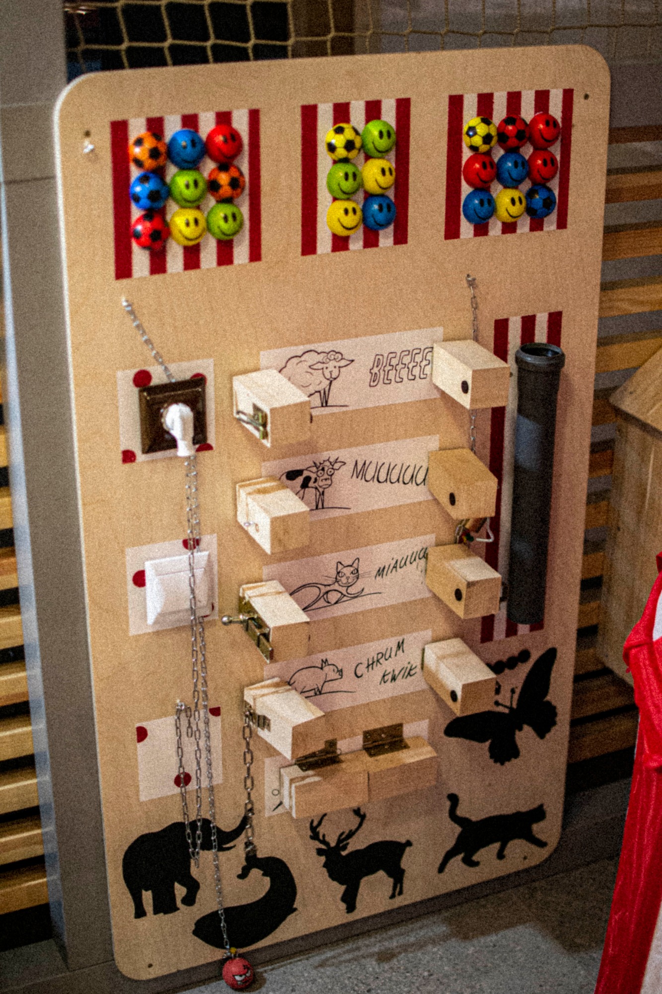 Drewniana tablica edukacyjna dla dzieci z elementami sensorycznymi: kolorowe piłki z emotikonami, włączniki światła, gniazdko, elementy z dźwiękami zwierząt oraz czarne sylwetki zwierząt na dole.