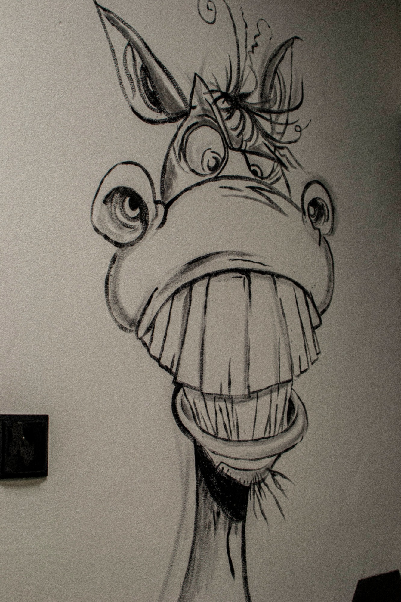 Szkic ołówkiem karykaturalnego konia z dużymi zębami na szarej ścianie, obok włącznika światła.