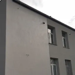 Szara elewacja budynku po renowacji, widoczne okna z białymi ramami, rynna i dwa punkty świetlne na ścianie.