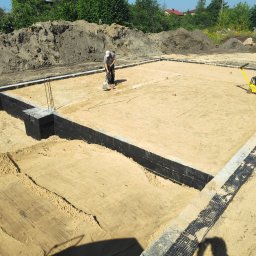 PUH RUFO Rafał Krupa - Fundamenty domu jednorodzinnego z izolacją pionową, wyrównany teren, pracownik z łopatą i zagęszczarka w tle.