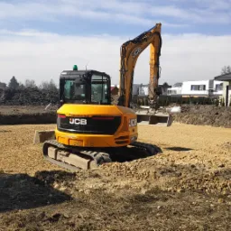 Żółta mini koparka JCB na placu budowy, przygotowany teren pod budowę domu, ubijarka w tle, widoczne fundamenty budynku.