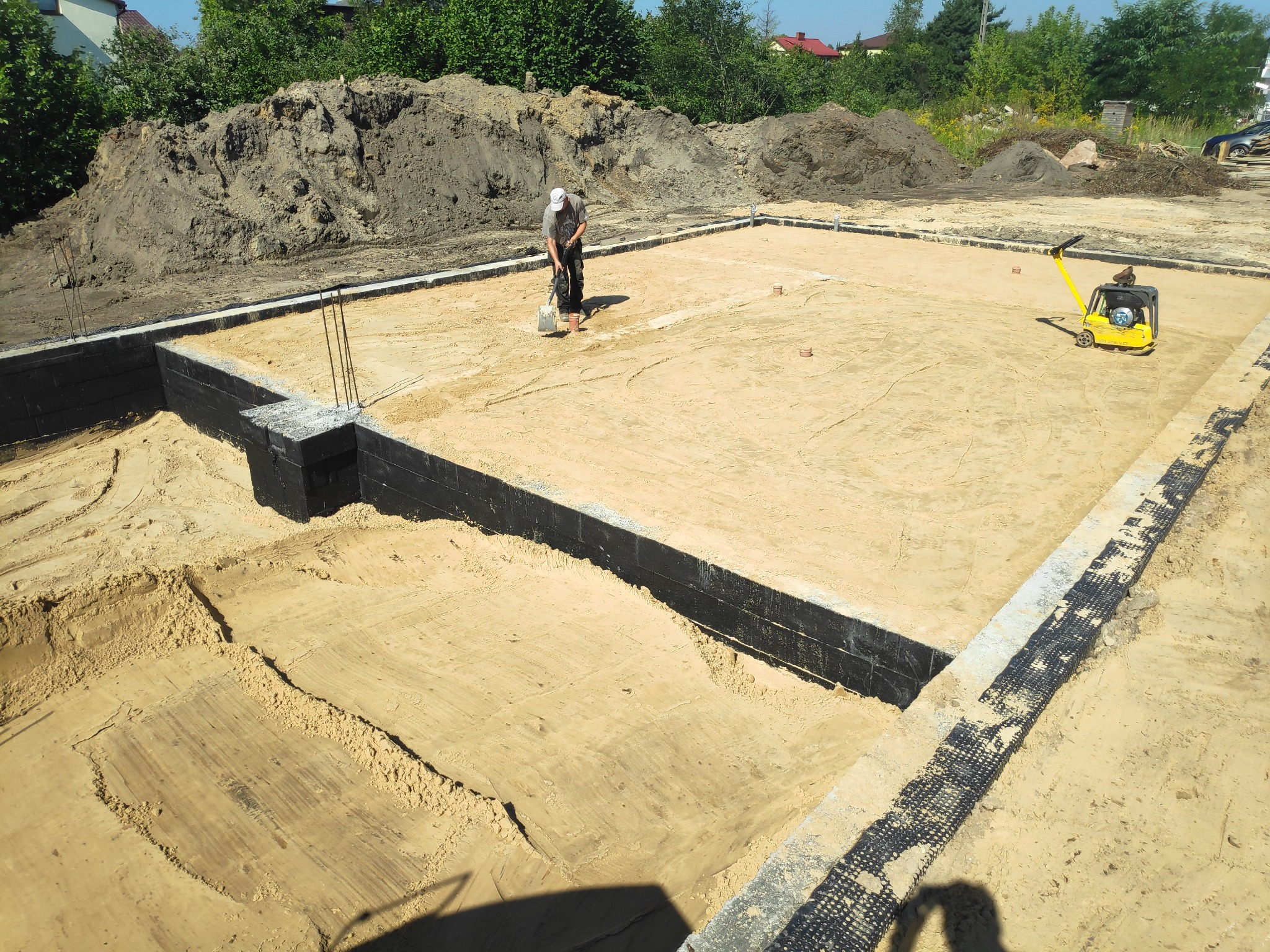 Fundamenty domu jednorodzinnego z izolacją pionową, wyrównany teren, pracownik z łopatą i zagęszczarka w tle.