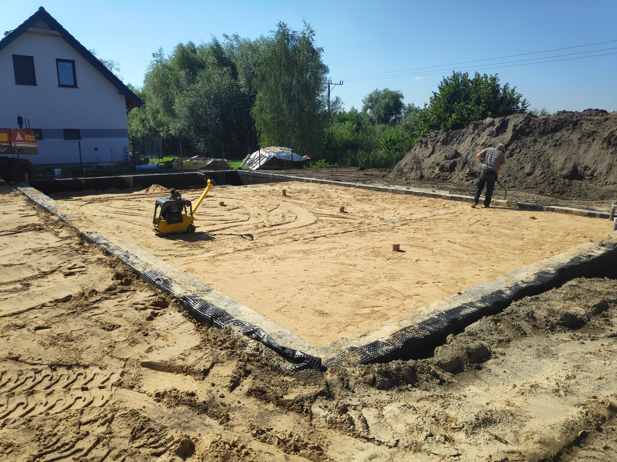 Przygotowany teren pod budowę domu jednorodzinnego; widoczne fundamenty, ubijarka do piasku, oraz pracownik wyrównujący teren.